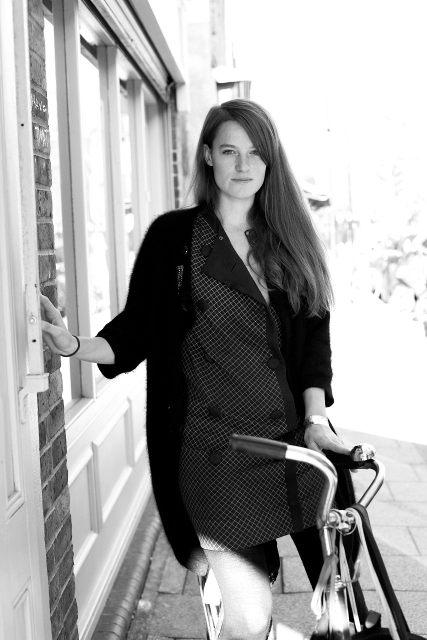 Black and white portrait of a woman with long dark hair standing by a window holding a bicycle.