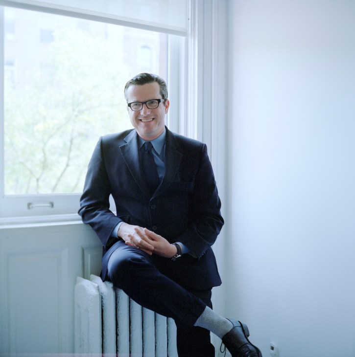 Man in dark suit and glasses sitting by a window in a bright interior space.