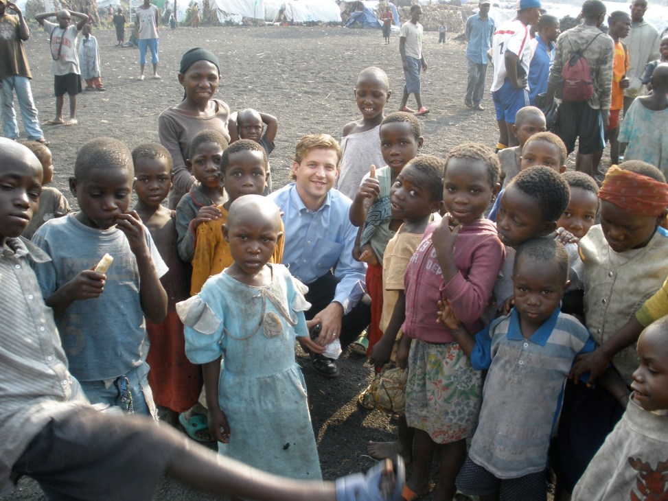 A man in a blue shirt surrounded by African children at a refugee camp.