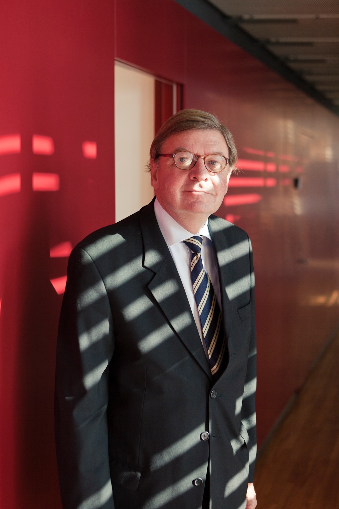 Man in dark suit and striped tie standing against red wall with window reflections.