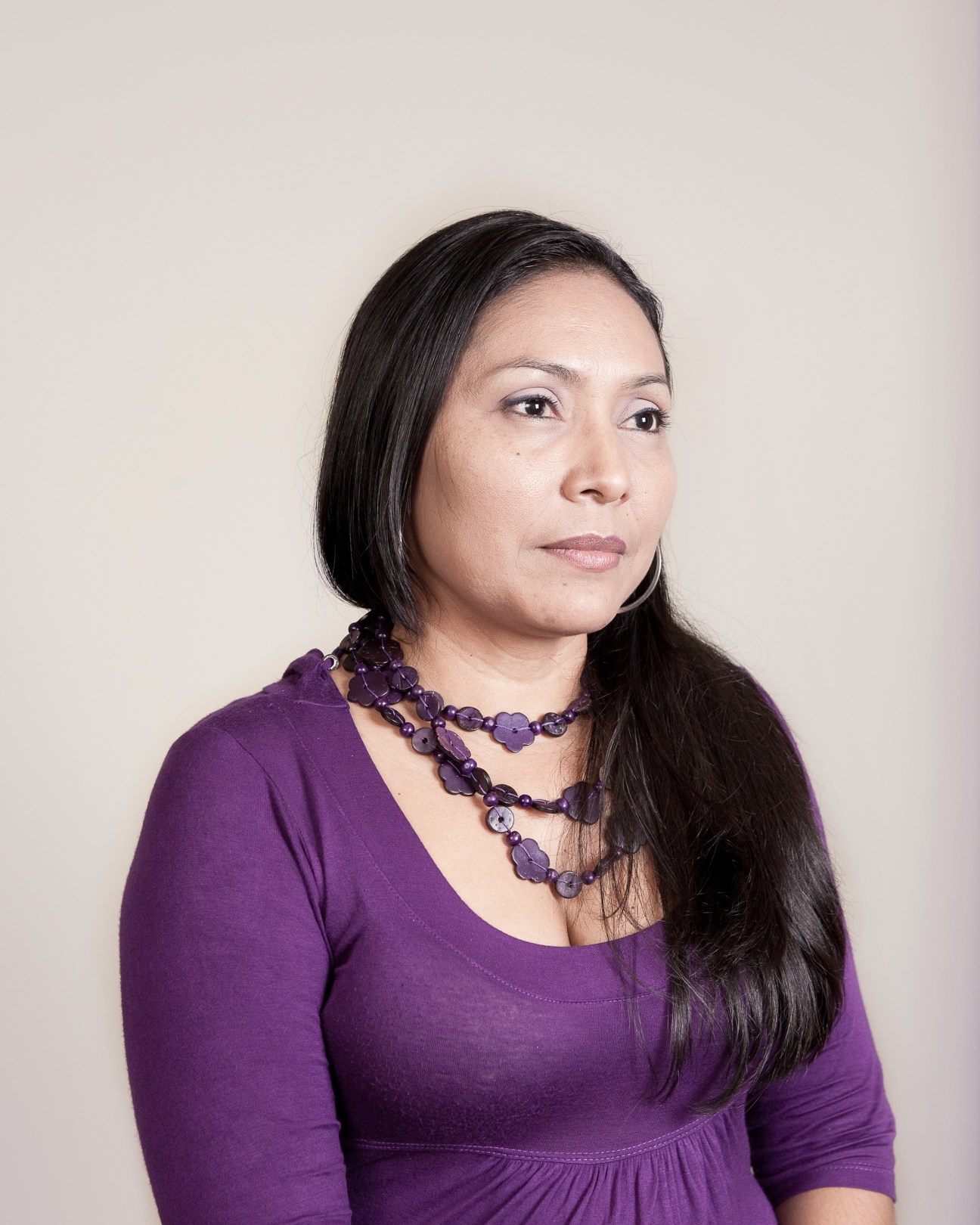 Woman wearing a purple top and beaded necklace against a neutral background.