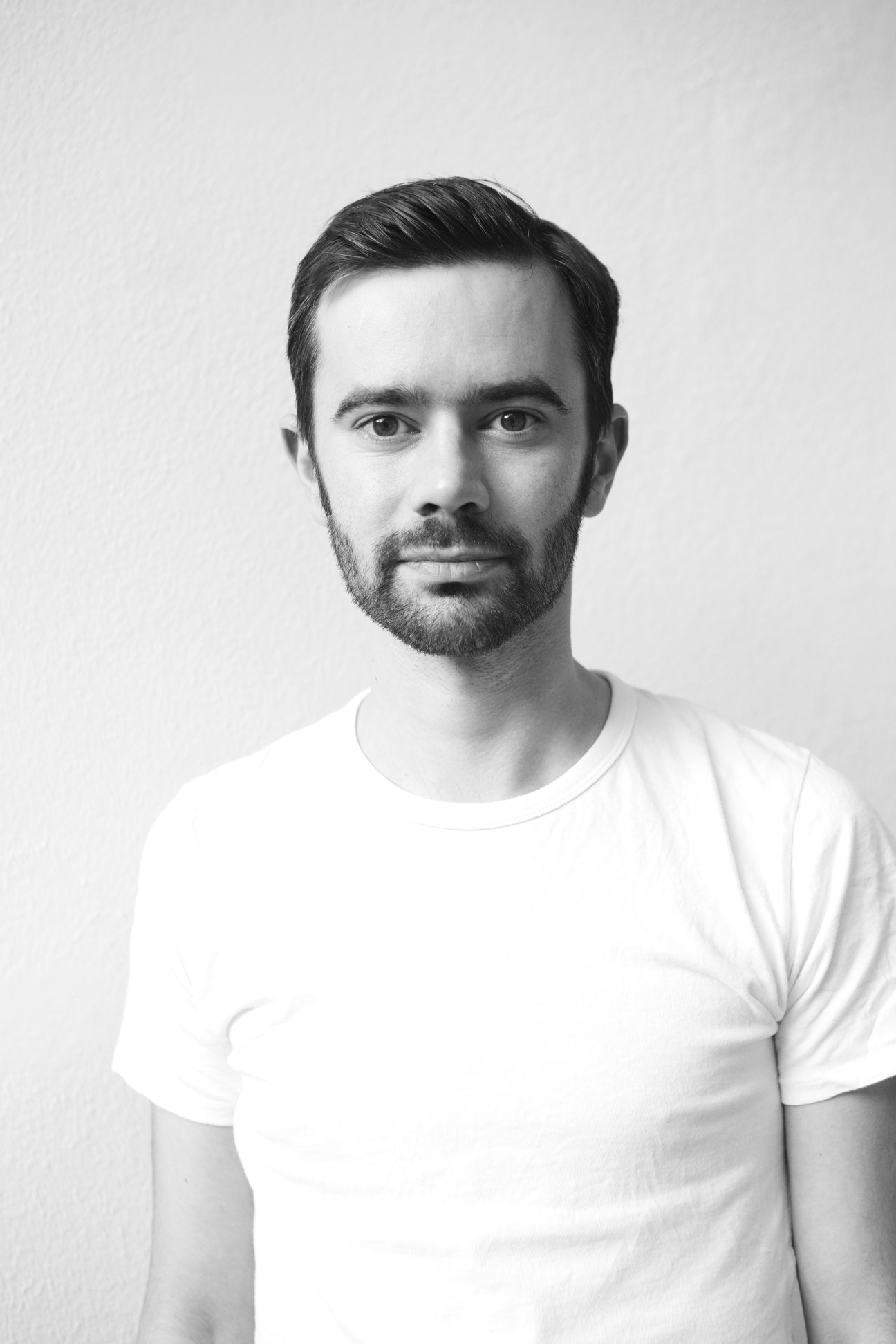 Black and white portrait photograph of a man with dark hair and beard wearing a white t-shirt.