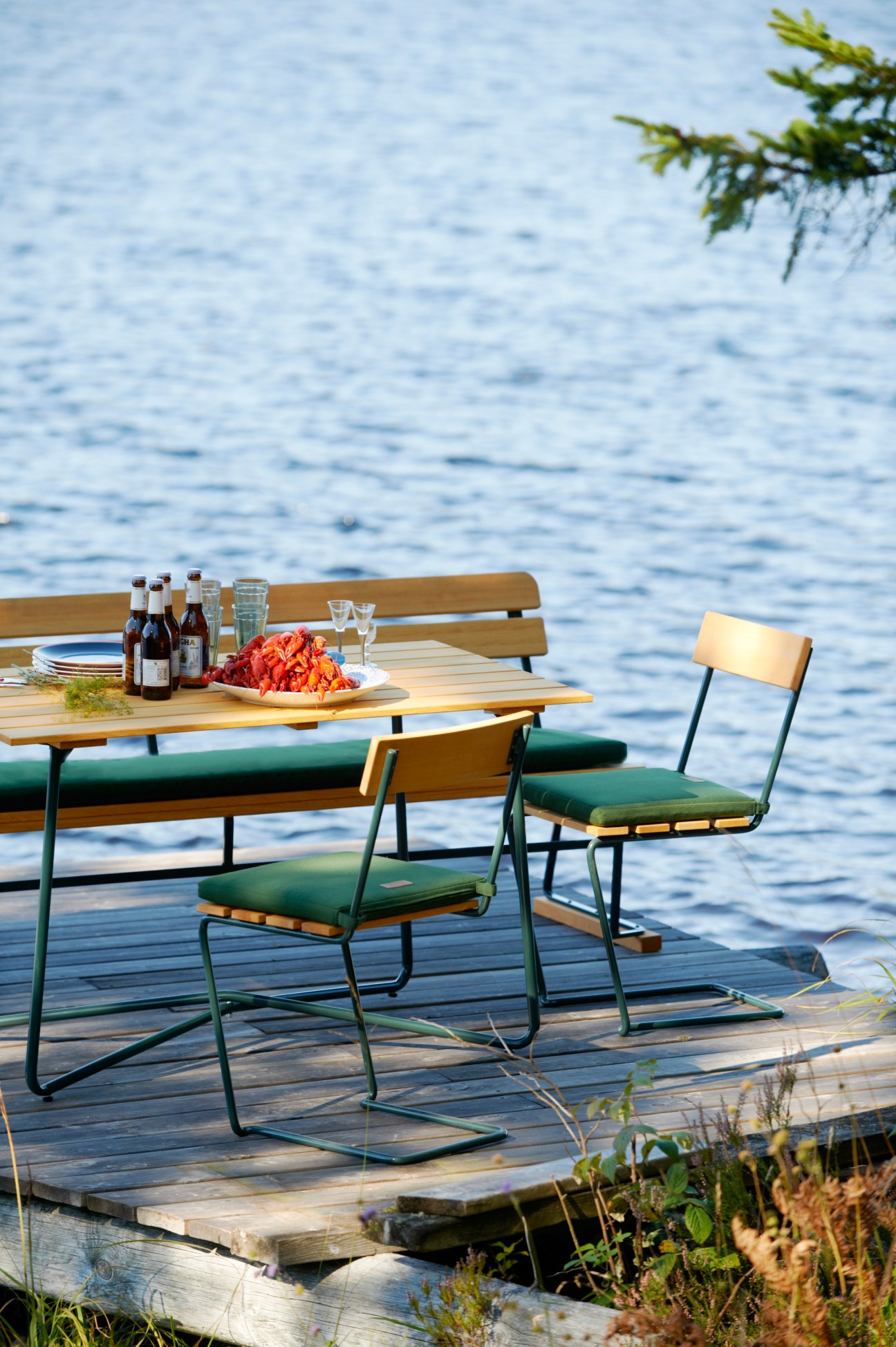 Outdoor dining set with wooden table and metal-framed chairs with green cushions on a lakeside deck.