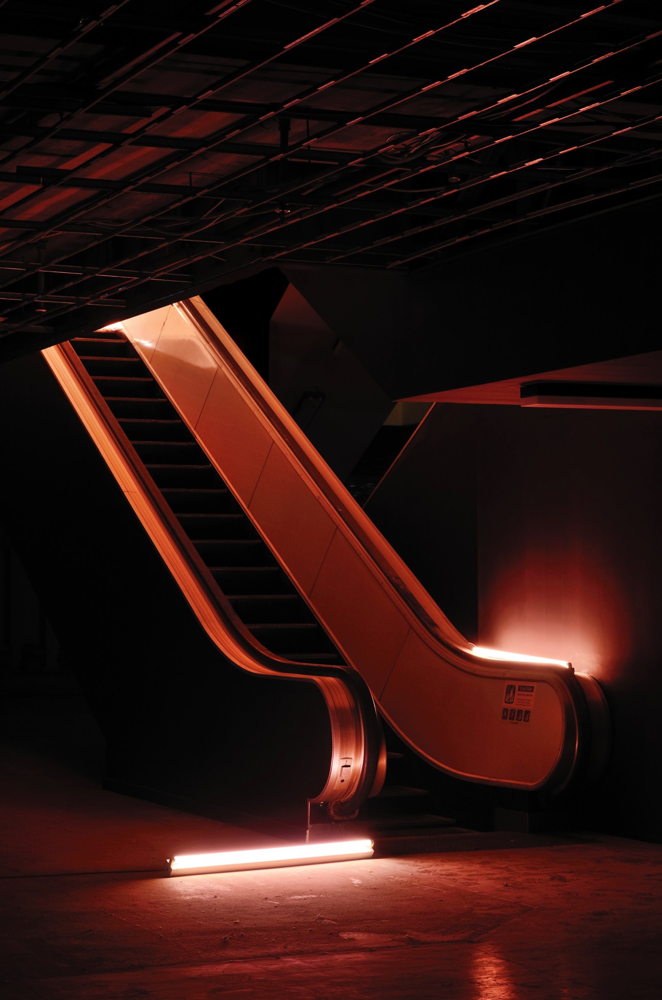 A large-scale red neon installation artwork glowing in a dimly lit gallery space with industrial ceiling.