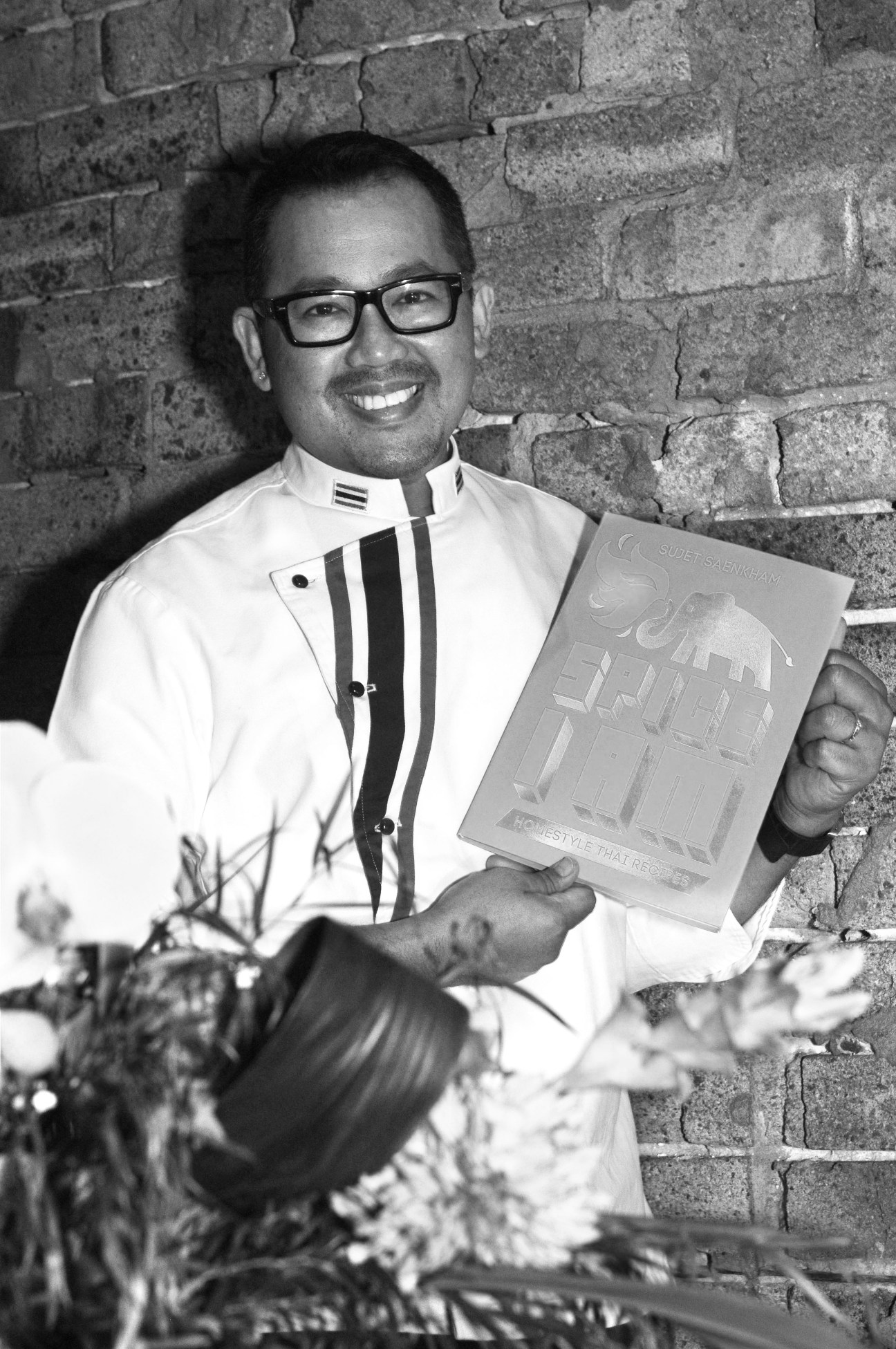 Sujet Saenkham, founder of Spice I Am, holding a menu in a white shirt against a brick wall.