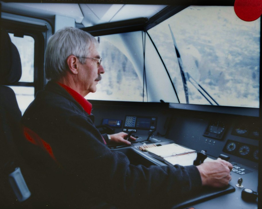 Passenger with white hair and glasses looks out window of Rhätische Bahn train cockpit at Alpine mountain landscape.