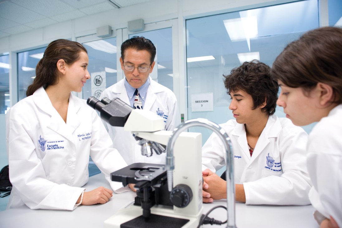 Medical students and instructor in white lab coats working with microscopes in a laboratory setting.