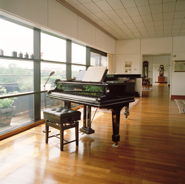 Grand piano in a modernist interior with polished wooden floors and floor-to-ceiling windows overlooking a courtyard.