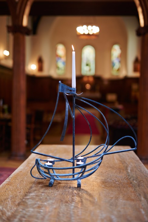 Ship-shaped iron candleholder with a lit candle on a wooden table in St Olav's chapel.