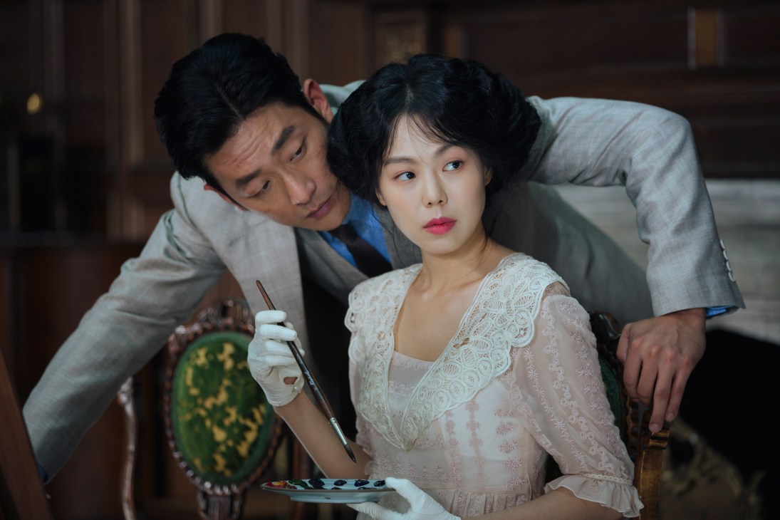 A woman in a cream lace dress holds a paintbrush while a man in a gray suit leans over her shoulder in a scene from The Handmaiden.