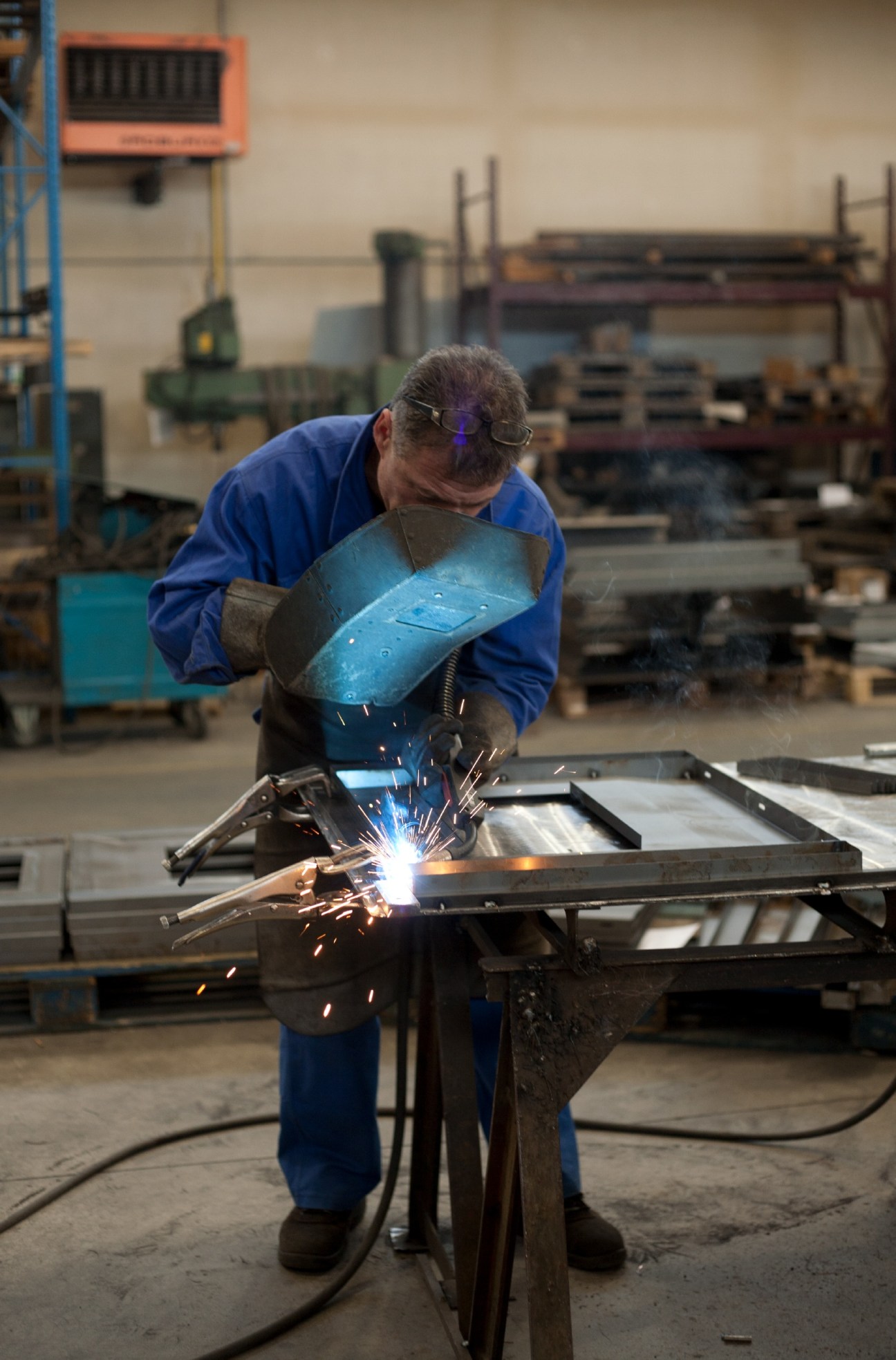 Welder in blue overalls and protective gear hand-welding steel safe components in factory.