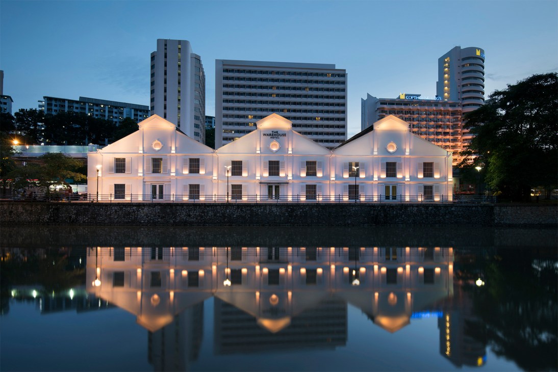 Warehouse Hotel illuminated at dusk, reflected in water, with modern buildings behind.