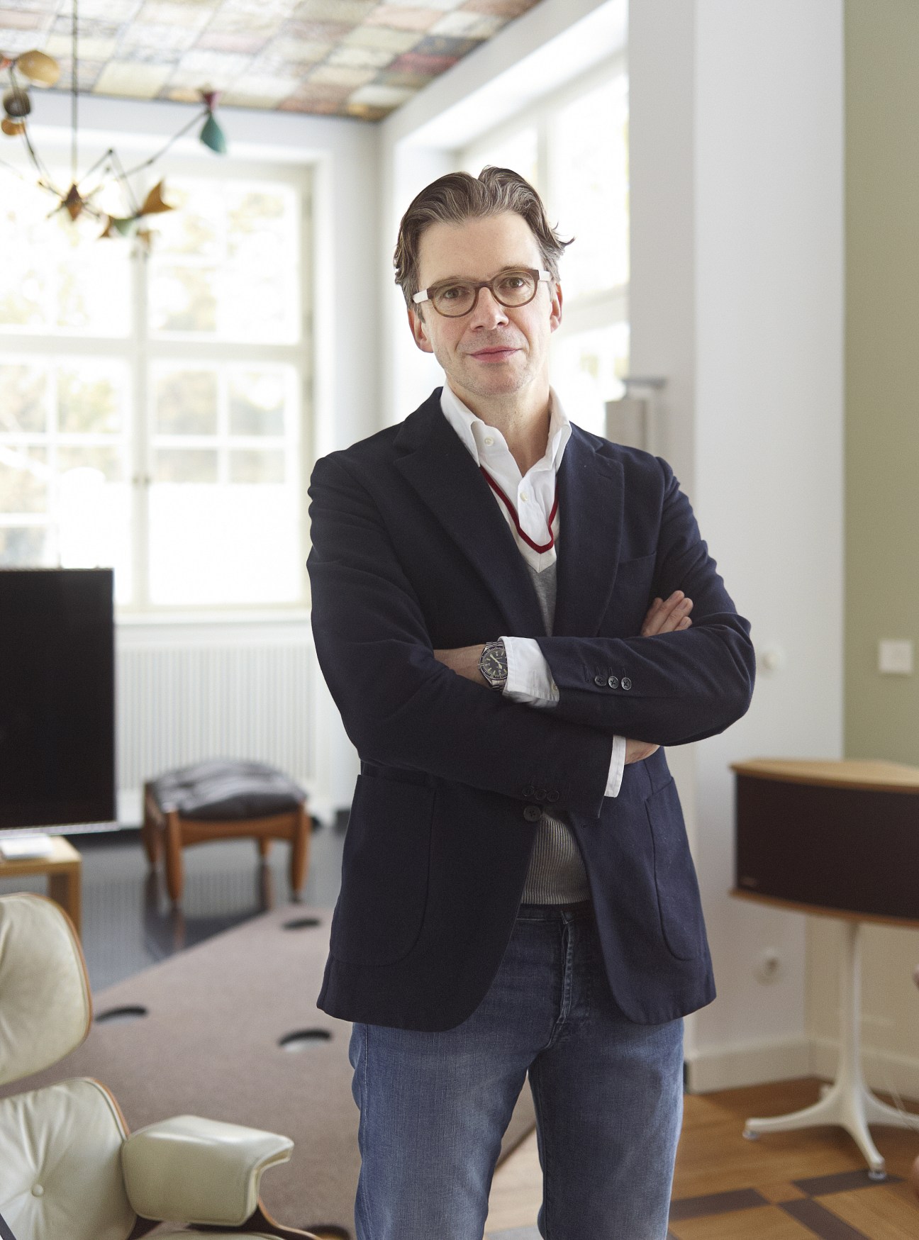 Thomas Andrae, a man wearing glasses and a dark blazer, stands with arms crossed in a bright office space with modern furniture.