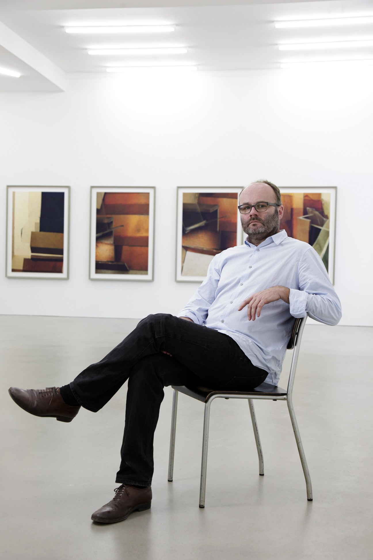 Artist Thomas Demand sitting in a gallery with framed photographs on the wall behind him.