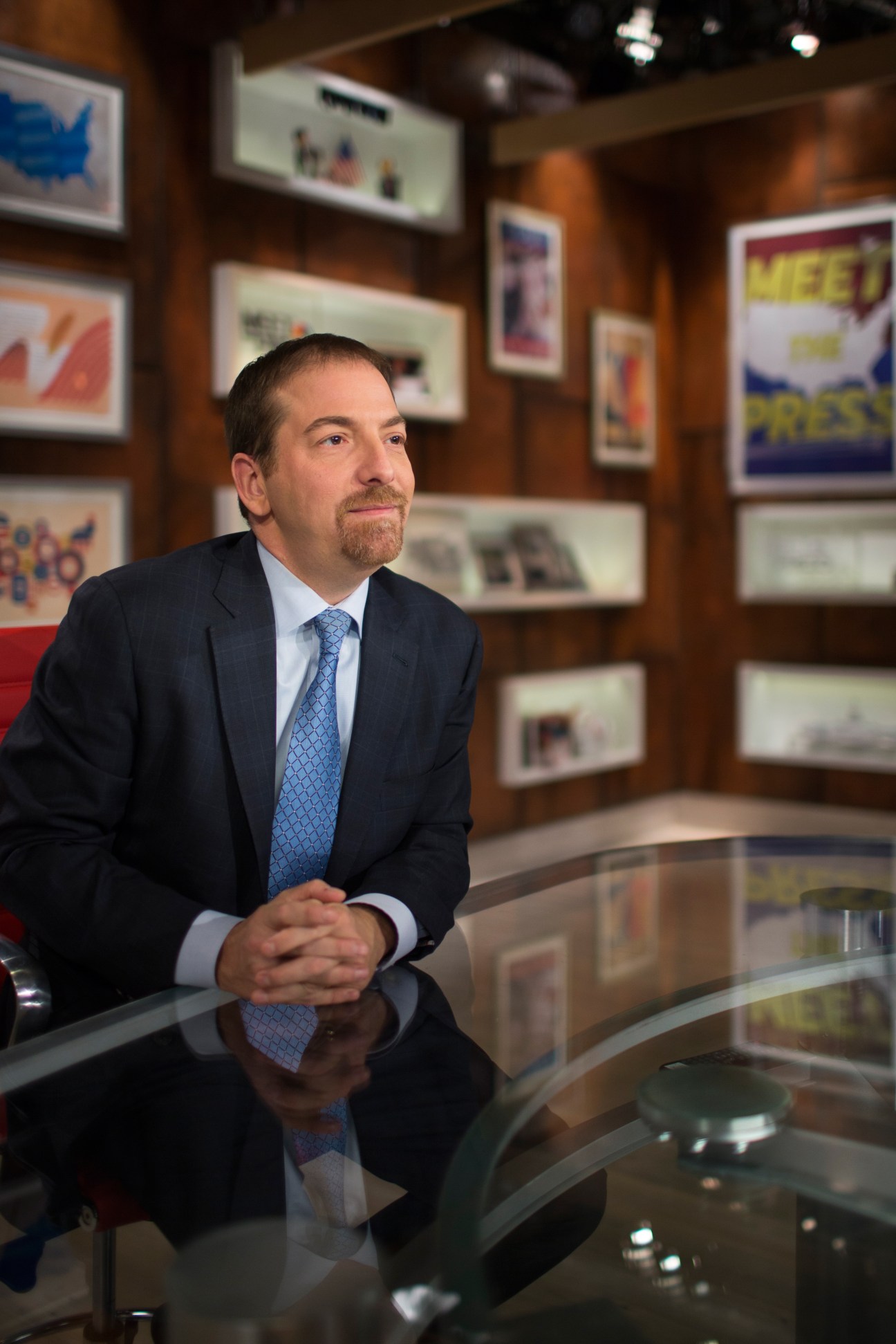 Chuck Todd sits at a desk in an NBC studio with television screens and framed images visible in the background.