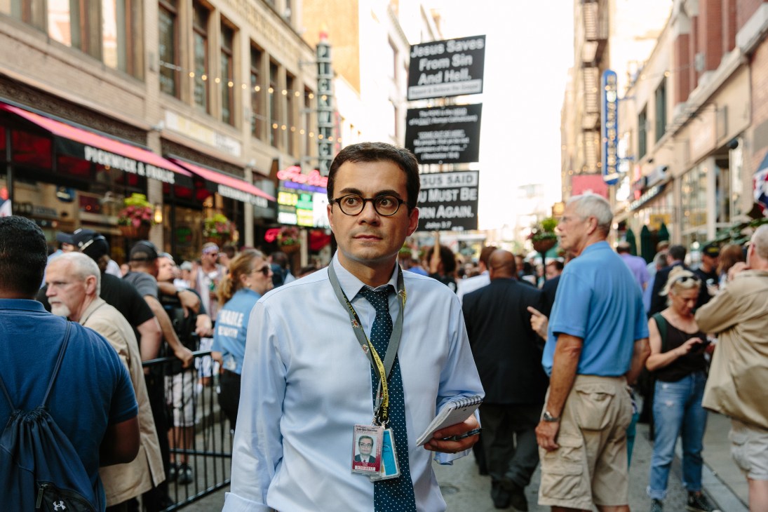 Tolga Tanis, a correspondent for Hurriyet, stands in Times Square wearing a press credential and holding a notebook amid campaign rally crowds.
