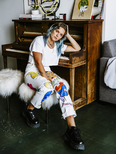 Swedish popstar Tove Lo sits on a stool in front of a wooden piano, wearing a white t-shirt and colorful patterned pants.