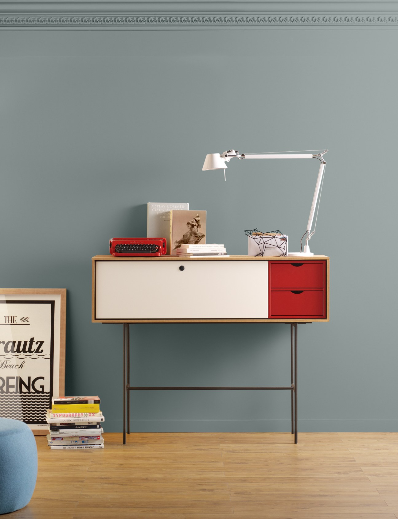Mid-century modern desk with white and red drawers, metal legs, topped with a red typewriter, books, and an adjustable desk lamp.