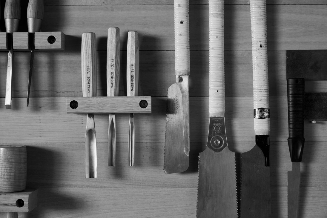 Black and white photograph of woodworking hand tools including chisels, saws, and files organized on a wooden wall-mounted rack.