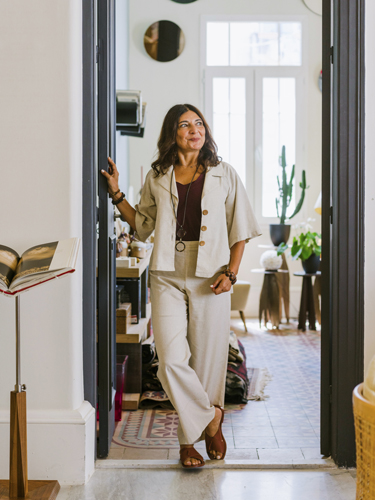 Woman in cream-colored suit standing in a bright hallway with plants and home decor visible.
