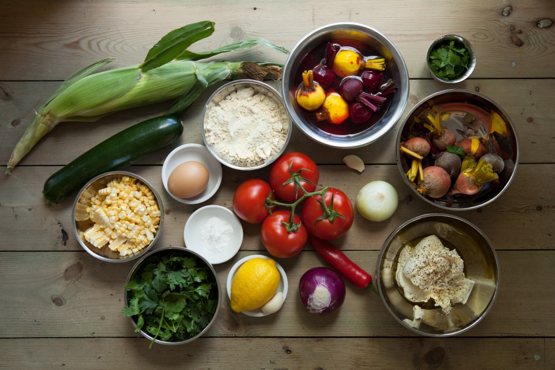 Fresh ingredients for sweetcorn and courgette fritters including corn, tomatoes, beetroot, herbs, and spices arranged on a wooden surface.