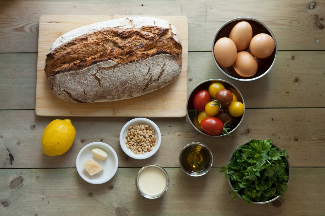 Sourdough bread, eggs, tomatoes, lemon, garlic, pine nuts, parmesan, parsley, and olive oil arranged on a wooden table.