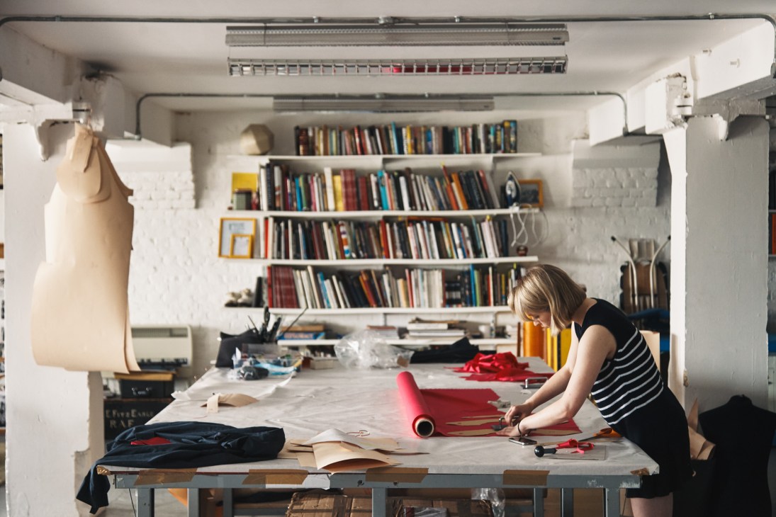 Designer working at a cutting table in E Tautz studio with fabric samples and reference books on shelves.