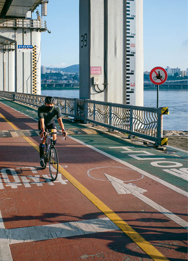 Cyclist riding on a colorful riverside path along the Han River in Seoul with bridges and mountains visible in the background.