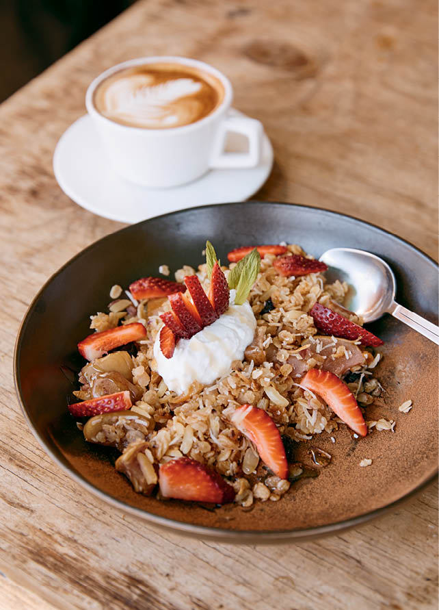 A bowl of granola with fresh strawberries and yoghurt alongside a latte on a wooden table.
