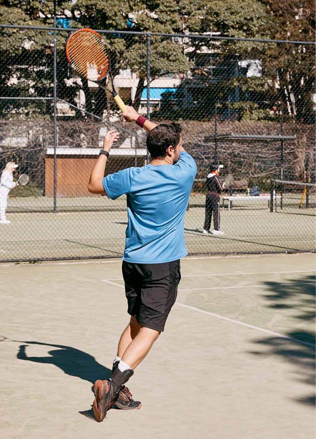 A man playing tennis at Rushcutters Bay Tennis Centre, serving a ball with a racket on a sunny court surrounded by trees.