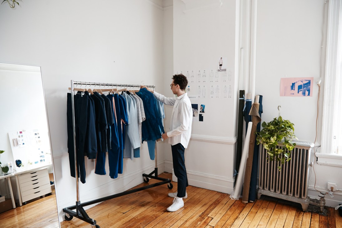 Designer in white shirt reviewing clothing on rack in bright studio space with wood floors.