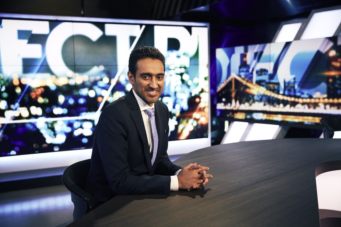 Waleed Aly seated at a desk on a television studio set with cityscape graphics behind him.