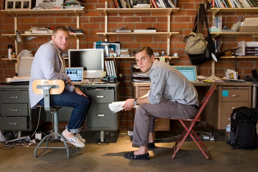 Two men in casual clothing sit in a loft office with exposed brick walls and wooden shelving, surrounded by computers and design materials.