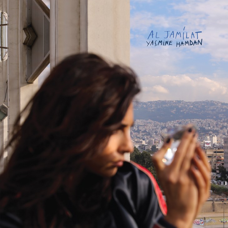 Woman holding phone with cityscape view and handwritten text reading 'Al Jamilat Yasmine Hamdan'