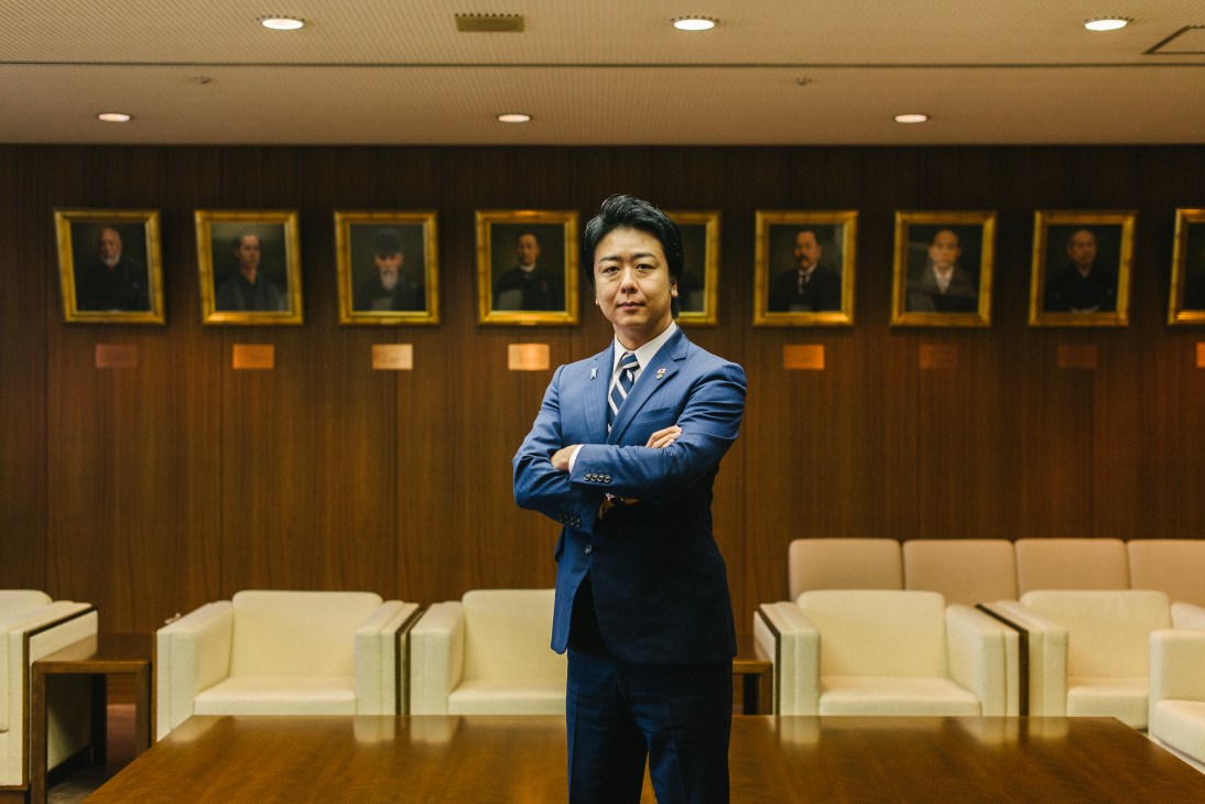 Soichiro Takashima, Mayor of Fukuoka, stands with arms crossed in a formal meeting room.