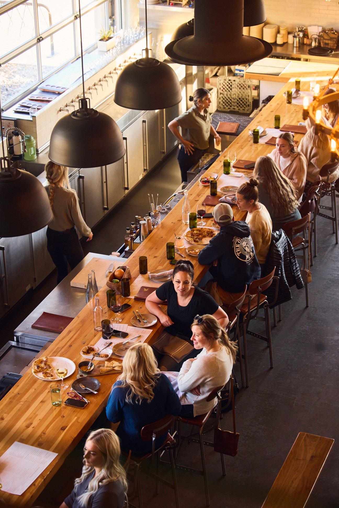 Overhead view of diners seated at long wooden table in modern restaurant with black pendant lamps.