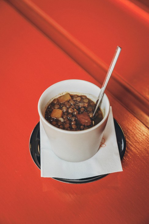 Bowl of lentil stew with beans and meat chunks served in white cup with spoon on black saucer