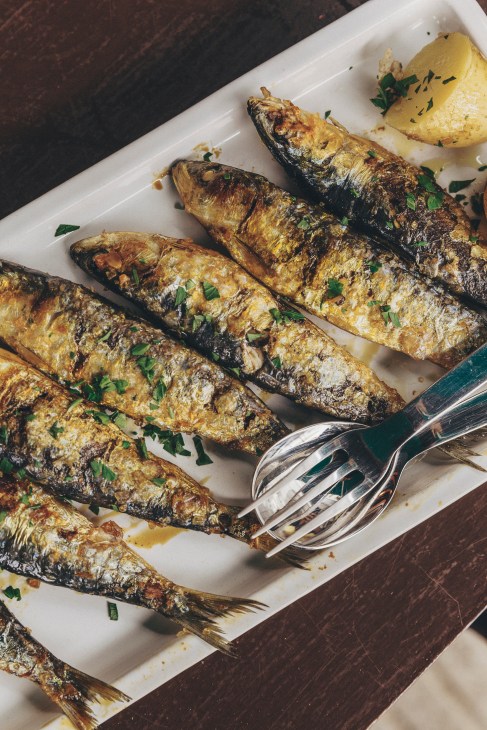 Grilled whole sardines garnished with herbs served on white parchment paper with lemon and fork