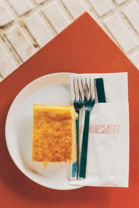 Slice of lemon cake on a white plate with fork and knife beside a napkin reading Bar do Porto