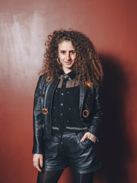 Sol wears a black leather jacket with decorative patches and matching leather shorts at La Paloma's carnival party
