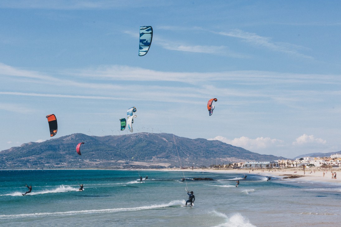 Multiple colorful kites flying above kitesurfers on Playa de los Lances beach in Tarifa with mountains in background
