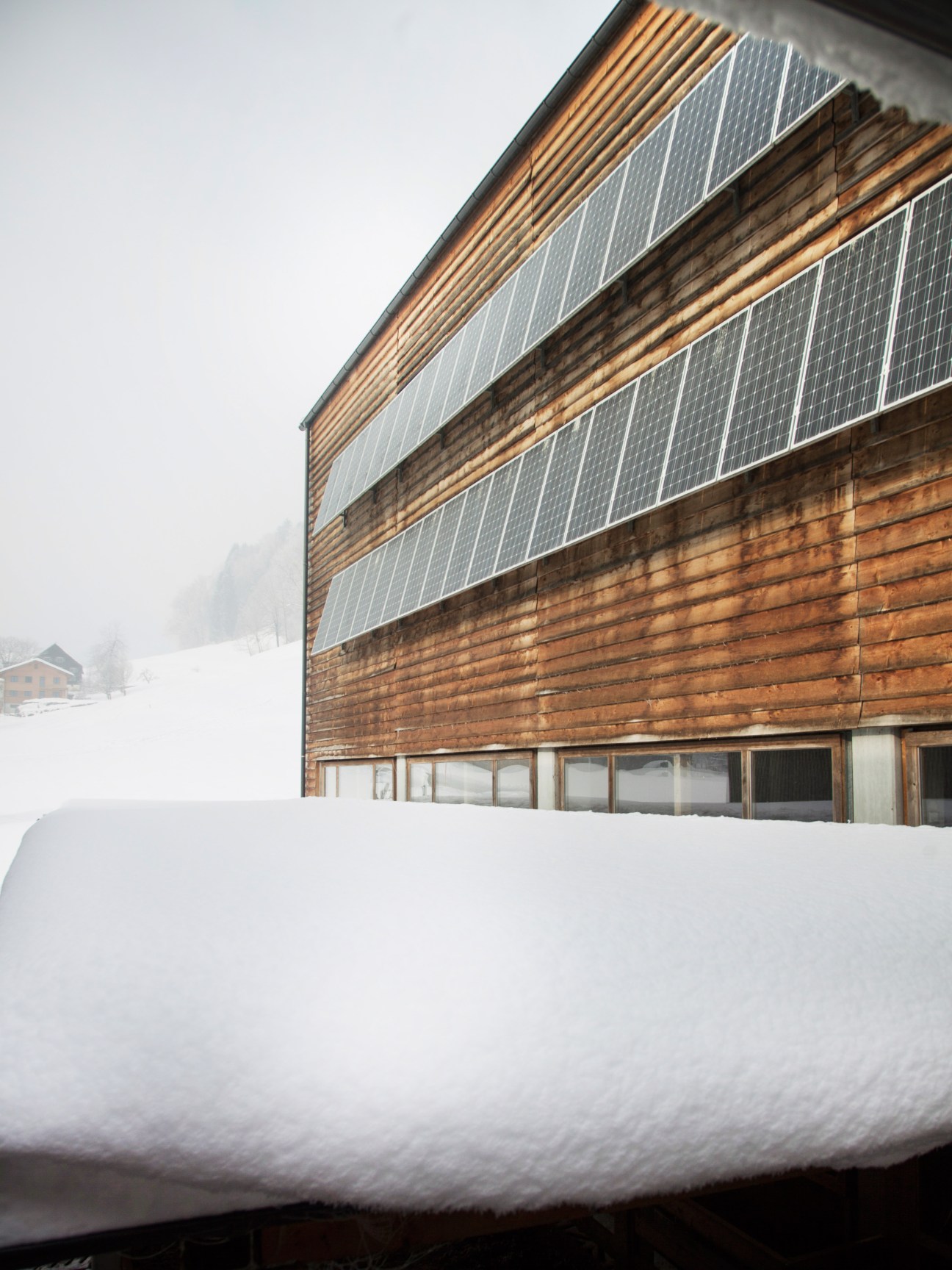 Modern wooden barn with solar panels mounted on facade, surrounded by snow in Austrian countryside.