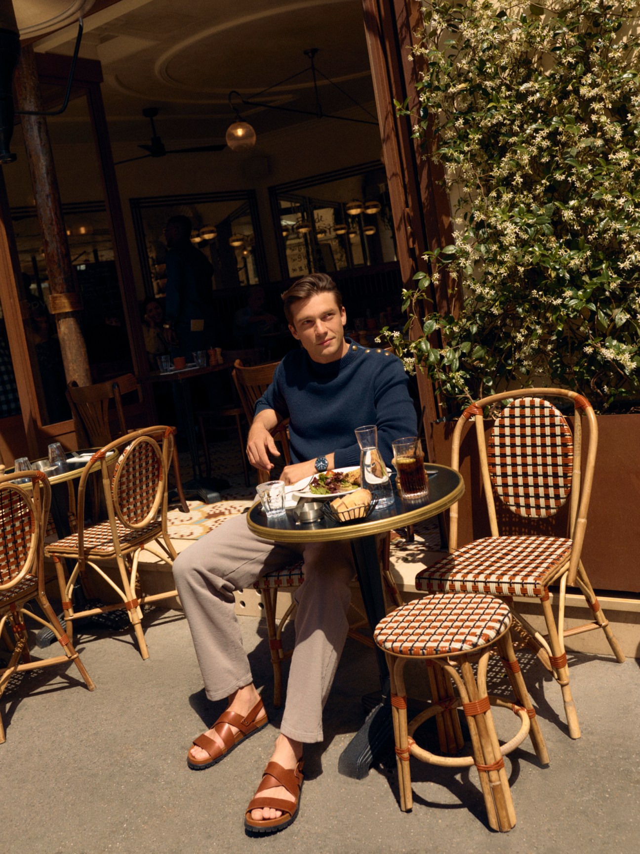 Man in blue sweater sitting at outdoor cafe table with wicker chairs near flowering tree