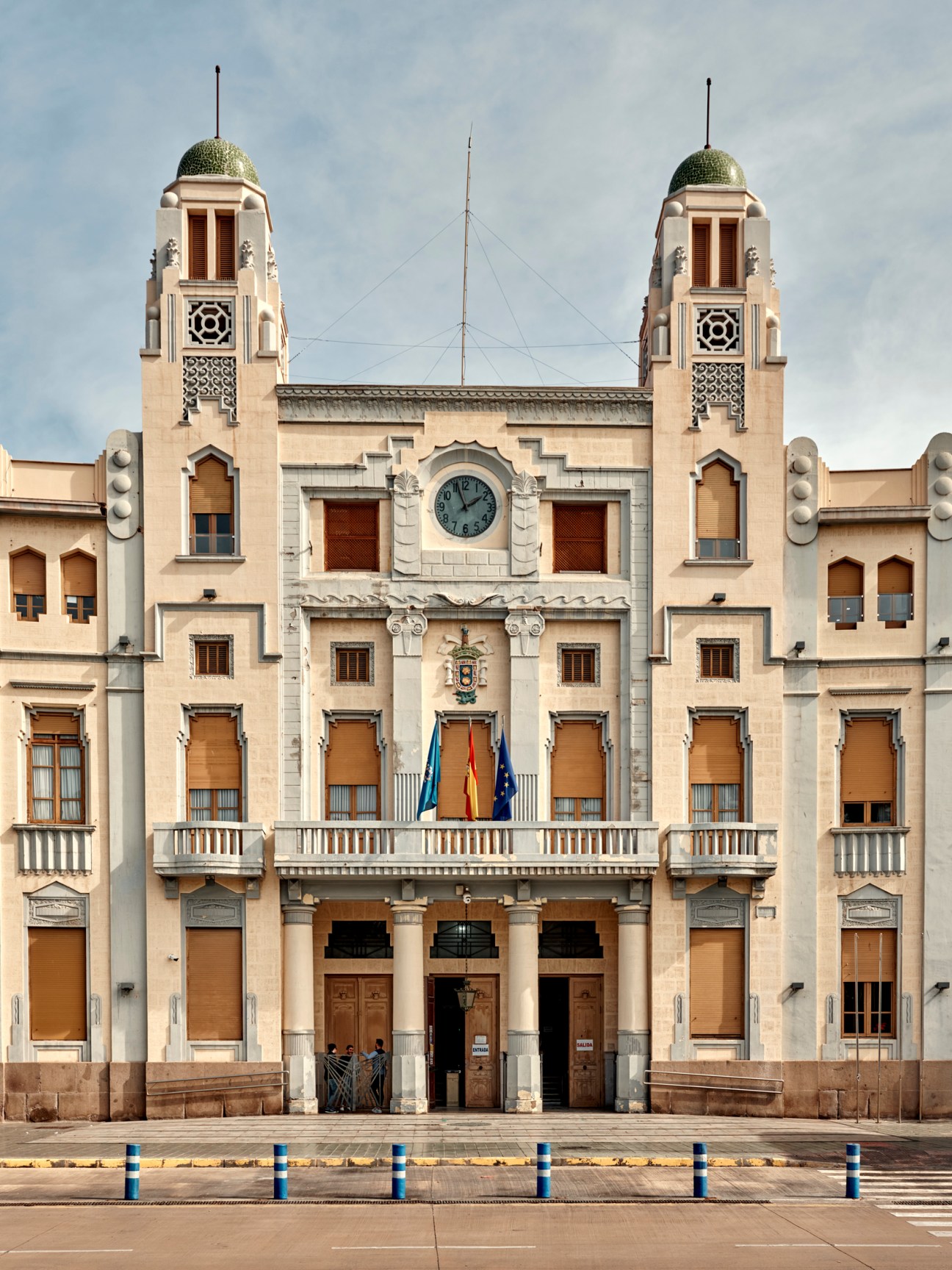 Symmetrical neoclassical city hall building with twin towers, green domes, clock, and Spanish flags on balcony