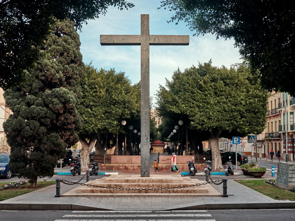 Stone cross monument on stepped platform surrounded by trees in central plaza with motorcycles parked nearby