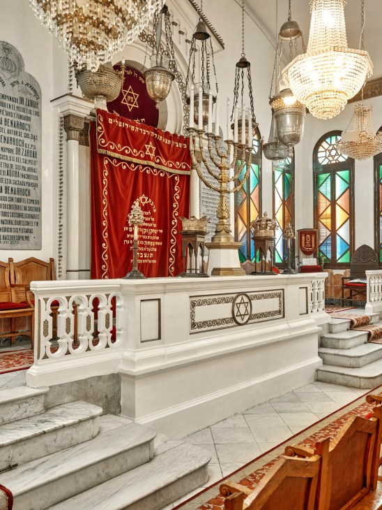 Interior of Or Zaruah Synagogue showing ornate chandeliers, red Torah ark curtain, and white marble bimah