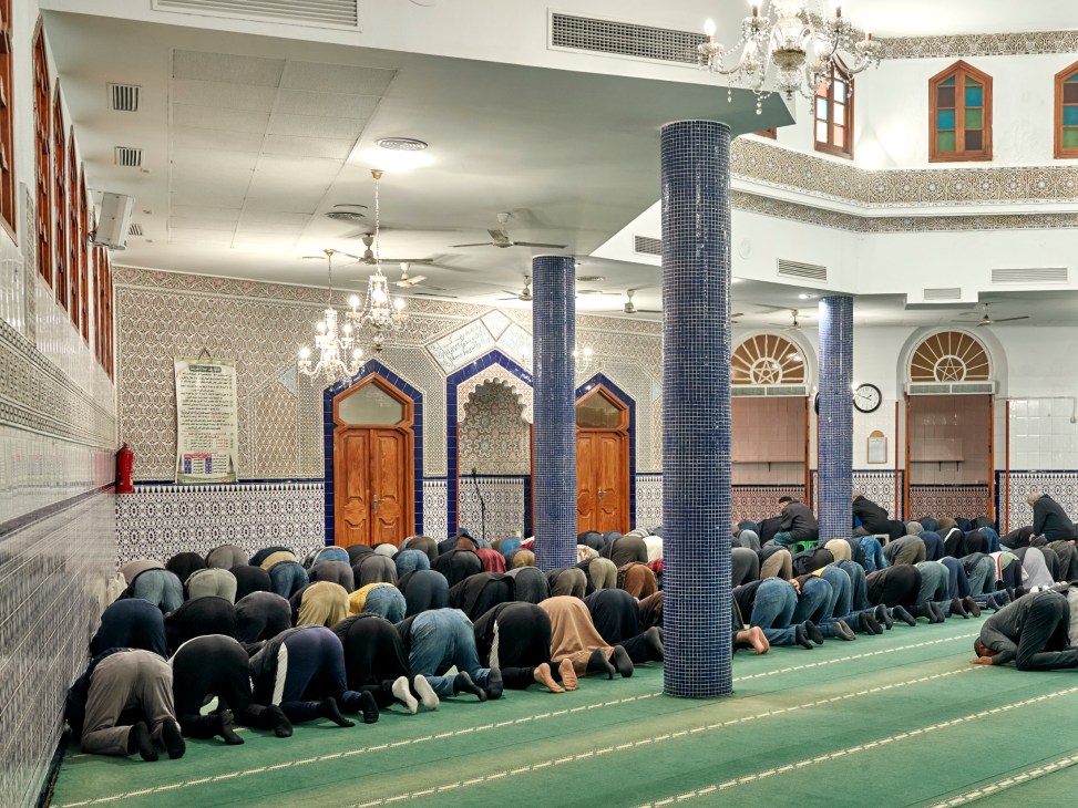 Worshippers prostrating in prayer on green carpeted floor of ornate mosque with blue tiled columns