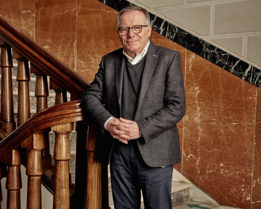 Eduardo de Castro in glasses and grey blazer stands beside an ornate wooden staircase in a formal marble-walled building