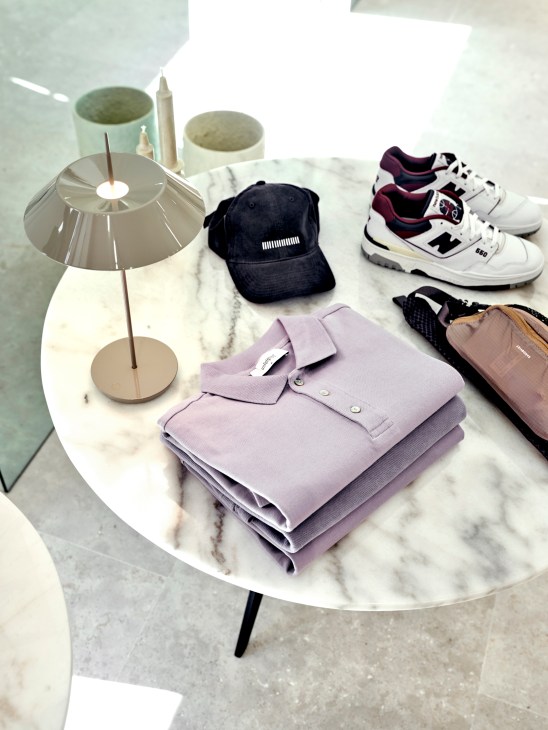 Folded lavender polo shirts, navy cap, white sneakers and small bag arranged on white marble table with modern lamp