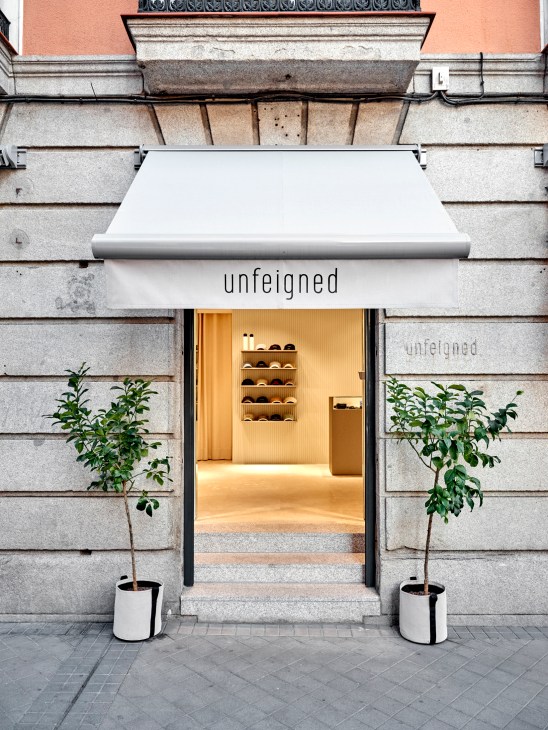 Modern storefront with white awning displaying 'unfeigned' brand name, warm interior lighting, and flanking planters